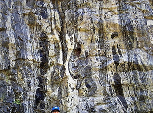 a rock structure and a hammer with red tape on the handle showing the scale of the different shapes of embedded boulders and layering