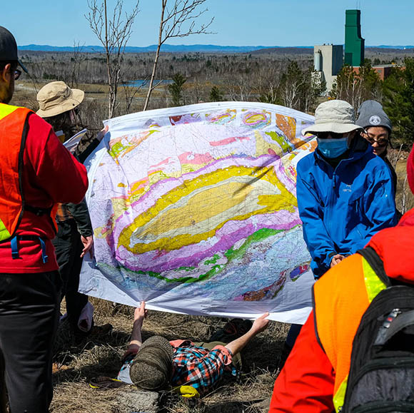 a colourful geologic map with students looking while a person lays on the ground holding the edge