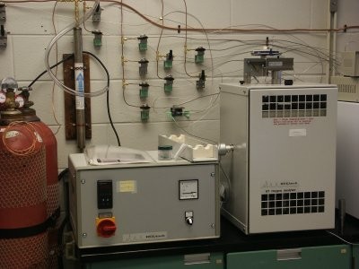 red pressurized gas tanks in a lab  next to two instruments on a green cabinet and a variety of plastic and metal tubing connections