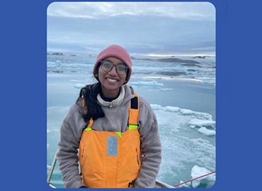 a female scientist in orange overalls standing on a ship with artic ice in the background