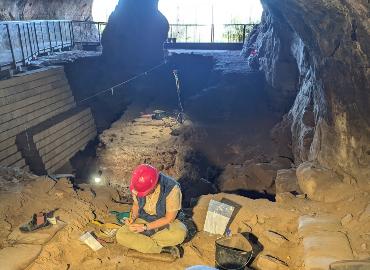 person sitting in a cave wearing a red hard hat and taking notes during an archaeology dig